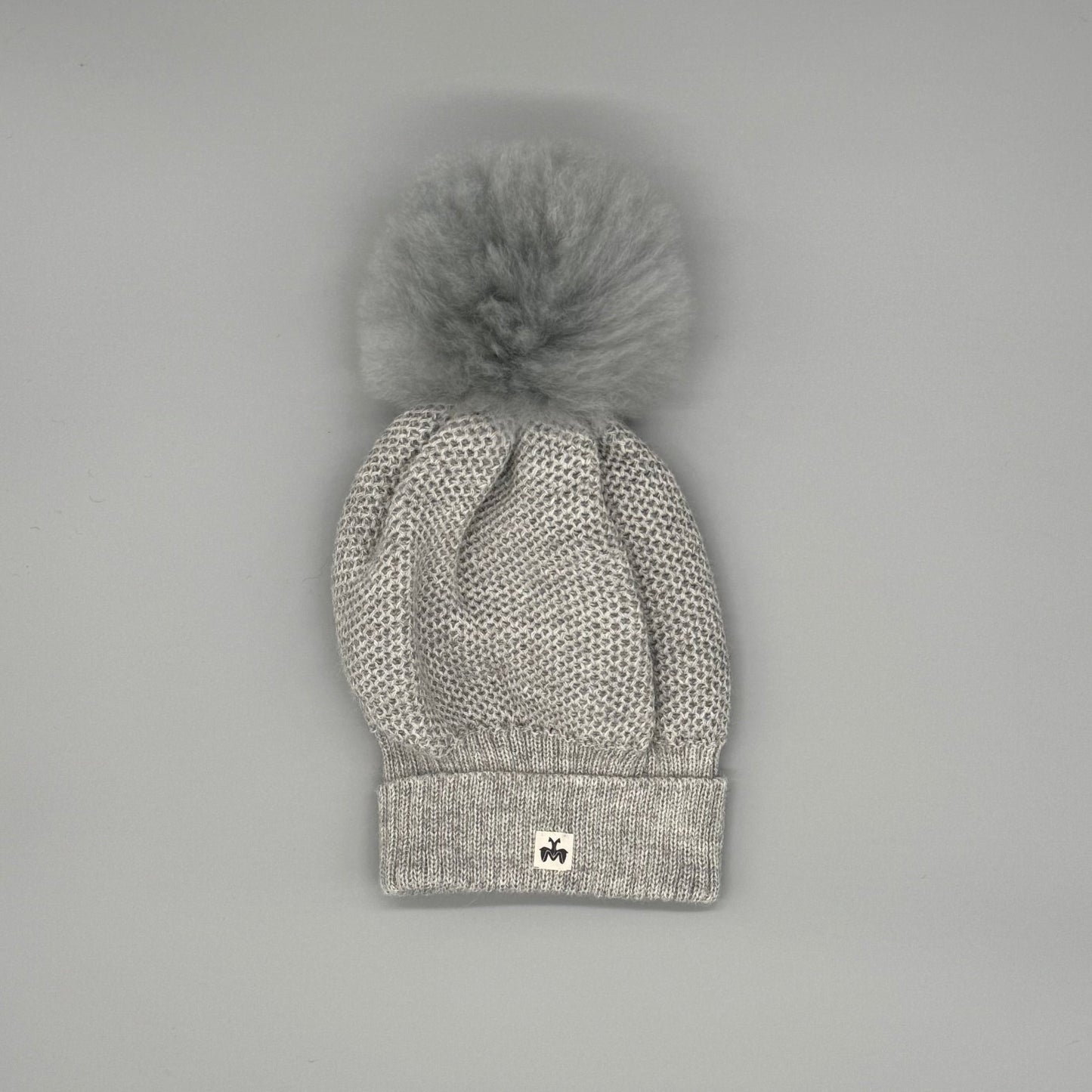 Alpakamütze Damen aus 100 % Babyalpaka in Grau mit Fellbommel, handgefertigt für stilvolle Wärme im Winter.