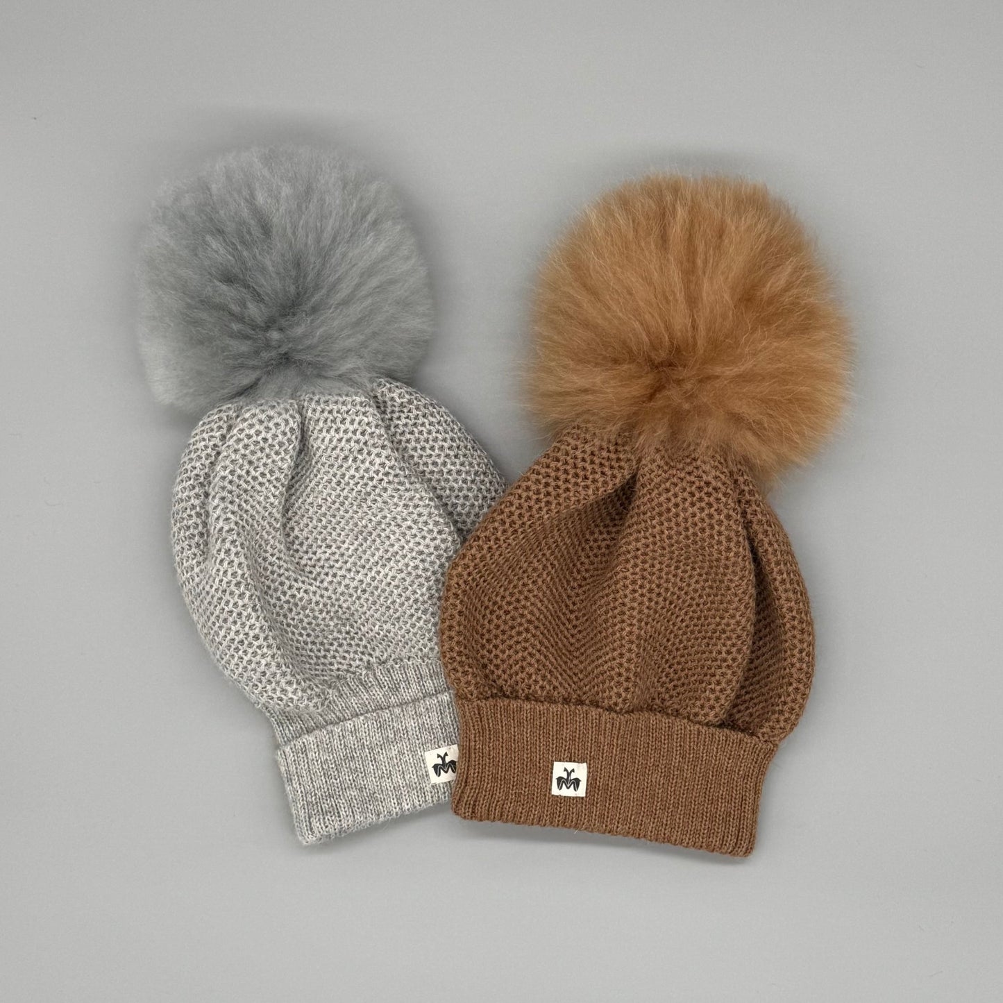 Zwei Alpakamütze Damen aus 100 % Babyalpaka in Grau und Braun mit Fellbommel auf grauem Hintergrund.